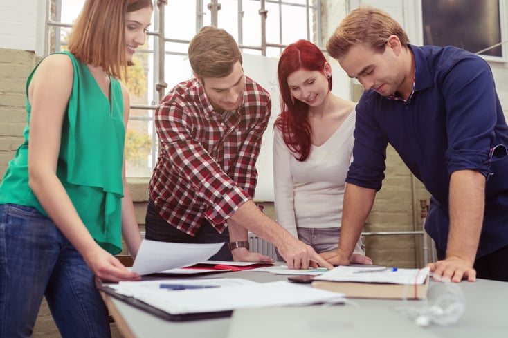 Group of Young White Friends Discussing the Project Paper at the Table While in Standing Position.