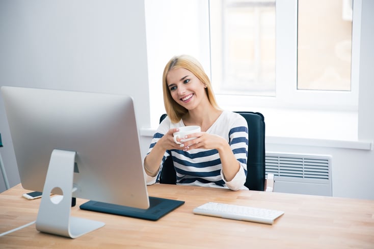 Happy female designer sitting at the table with desktop and drinking coffee in office