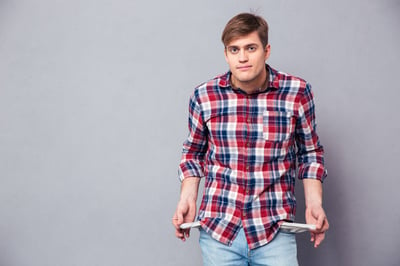 Poor handsome young man in checkered shirt and jeans showing empty pockets over grey background