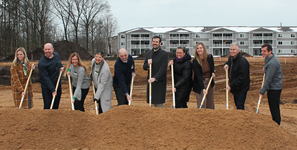 Group of people holding golden shovels at groundbreaking ceremony in front of residential apartment buildings on brown field