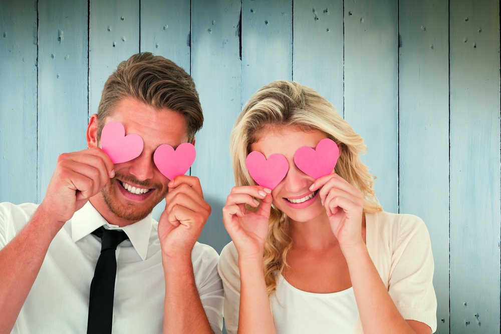 Attractive young couple holding pink hearts over eyes against wooden planks.jpeg