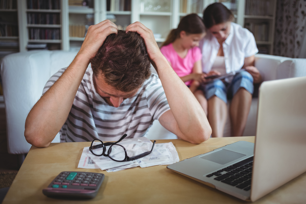 Worried man sitting at table with bills and laptop while his wife and daughter sitting on sofa